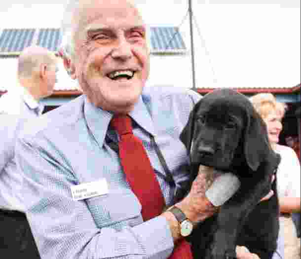 A Guide Dog's staff member holding a black eight week labrador puppy. The staff member is smiling at the camera.