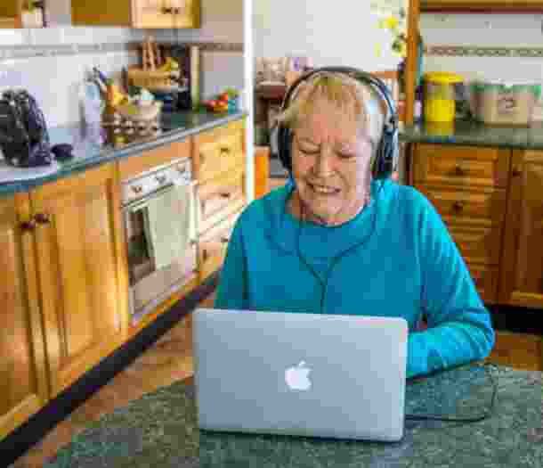 An older adult wearing headphones whilst on the laptop. The person is seated at a kitchen table.