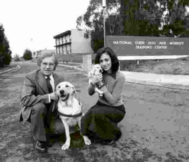 A 1940's black and white image of two people outside a Guide Dog Training Centre. One of the people in kneeling down next to a yellow labrador Guide Dog in harness and the other is holding a yellow ten week old labrador puppy. Both dogs and people are looking at the camera.