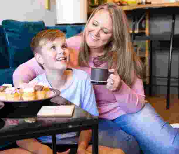 Mother sitting on the ground next to her child and they are both smiling. The mother is holding a mug and there is a plate of muffins on the table in front of them.
