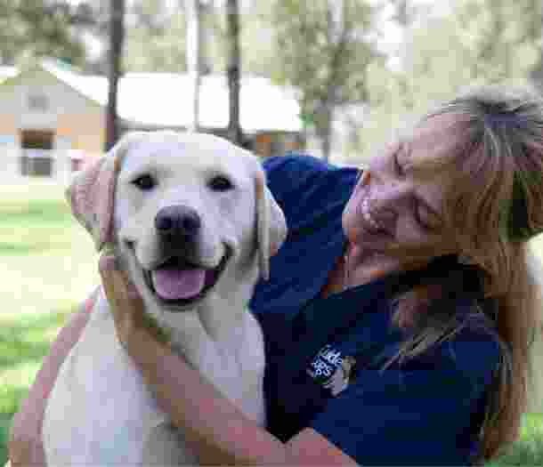 A veterinarian and fully grown yellow labrador are sitting outside. The veterinarian is looking at the dog with a smile on their face and the dog is smiling at the camera.