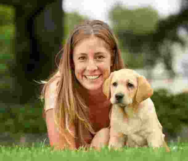 A person laying on some grass with an eight week old yellow labrador puppy. The person is smiling and looking at the camera. The puppy is also looking at the camera.