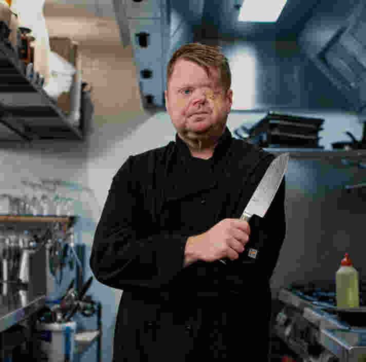 Craig stands in a commercial kitchen holding a large chef's knife.